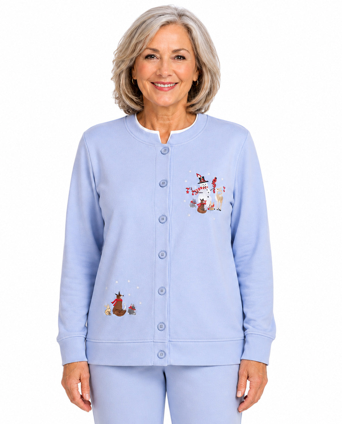 A smiling older woman wearing a light blue button-front cardigan and matching pants with embroidered holiday motifs, photographed against a plain white studio background.