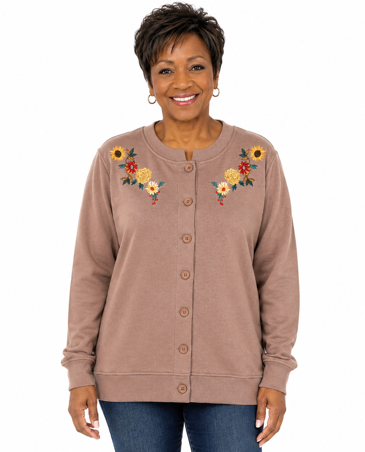 A woman wearing a taupe button-up cardigan with colorful floral embroidery on the chest, paired with blue jeans, against a plain white background.