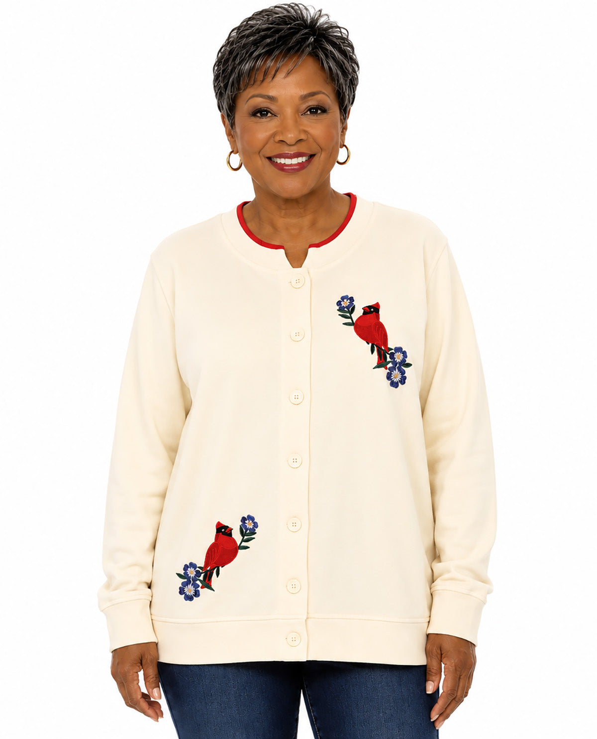 A smiling woman wearing a cream-colored button-up cardigan with red bird and blue flower embroidery, paired with dark blue jeans, against a plain white background.