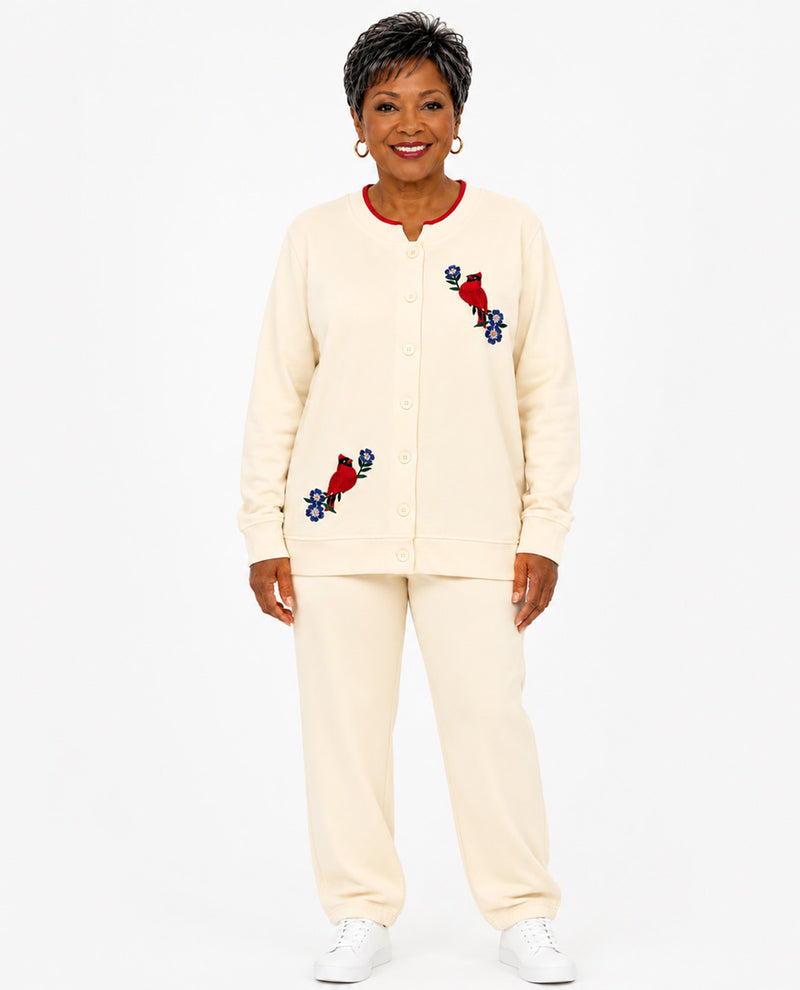 An older woman modeling a cream-colored two-piece lounge set consisting of a button-front cardigan and matching sweatpants, featuring red and blue embroidered birds on the chest and sleeves, set against a plain white background.