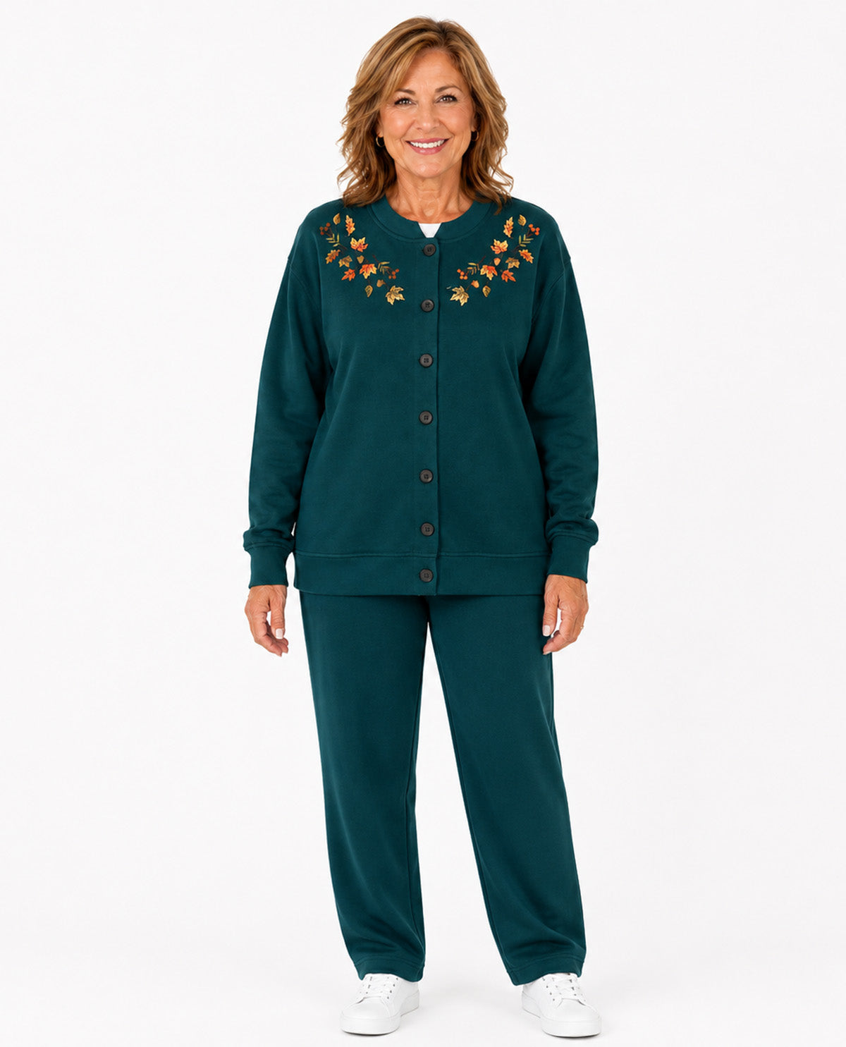 A smiling woman stands against a light gray studio background wearing a dark teal matching lounge set consisting of a button-front cardigan top with orange floral embroidery around the neckline and matching wide-leg pants.