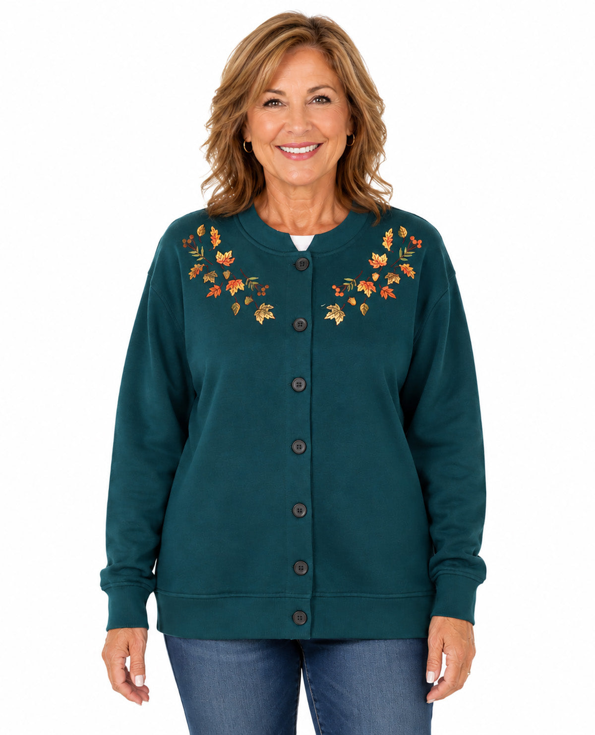 A smiling woman wearing a dark teal button-up cardigan sweater with embroidered autumn leaves in orange, yellow, and green on the chest, paired with blue jeans, against a plain white background.