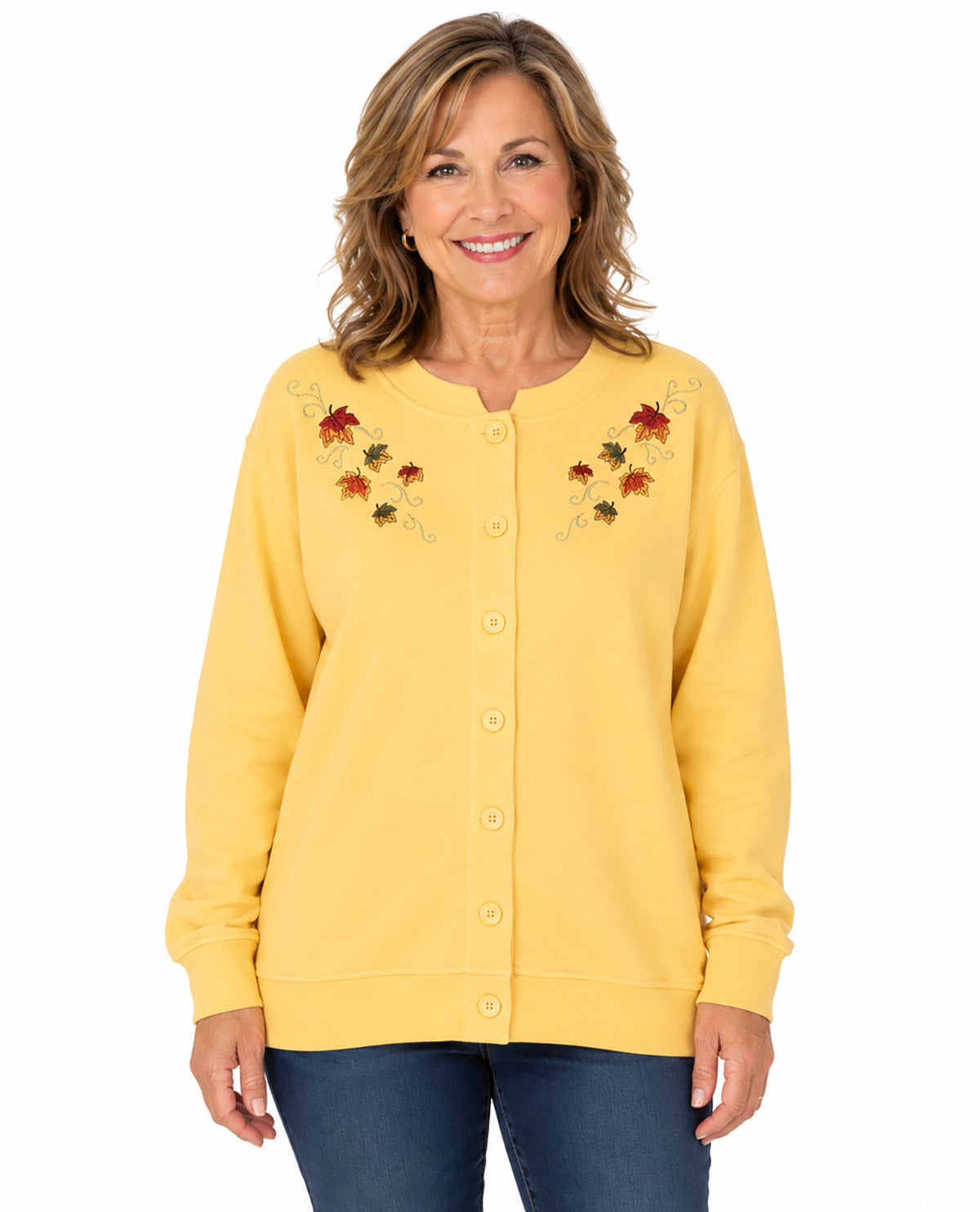 A smiling woman wearing a yellow button-front cardigan with embroidered autumn leaves on the chest, paired with dark blue jeans, against a plain white background.