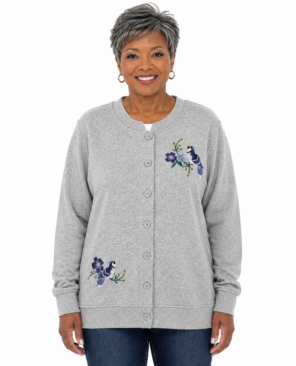 A smiling woman with short gray hair wearing a light heather gray button-up cardigan with purple and blue floral embroidery, paired with dark blue jeans, against a plain white background.