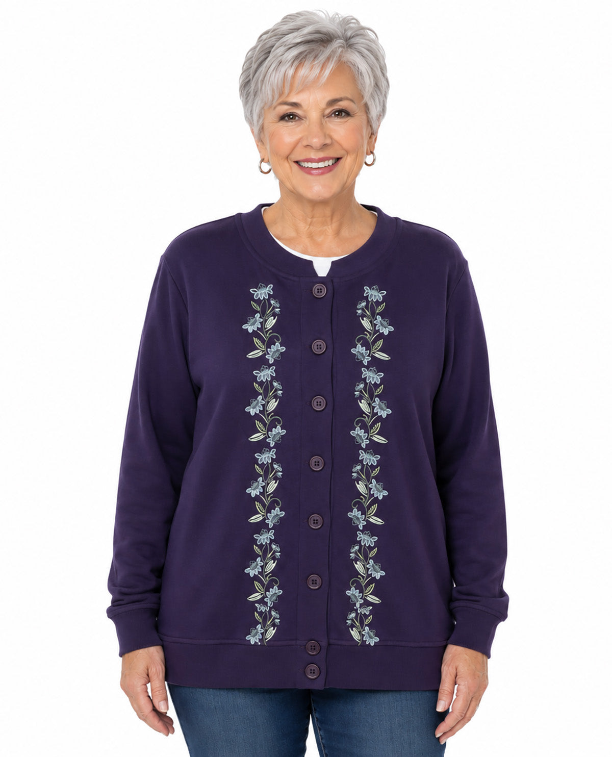 A smiling woman with short gray hair wearing a dark purple button-up cardigan with vertical floral embroidery down the front, paired with blue jeans, against a plain white background.