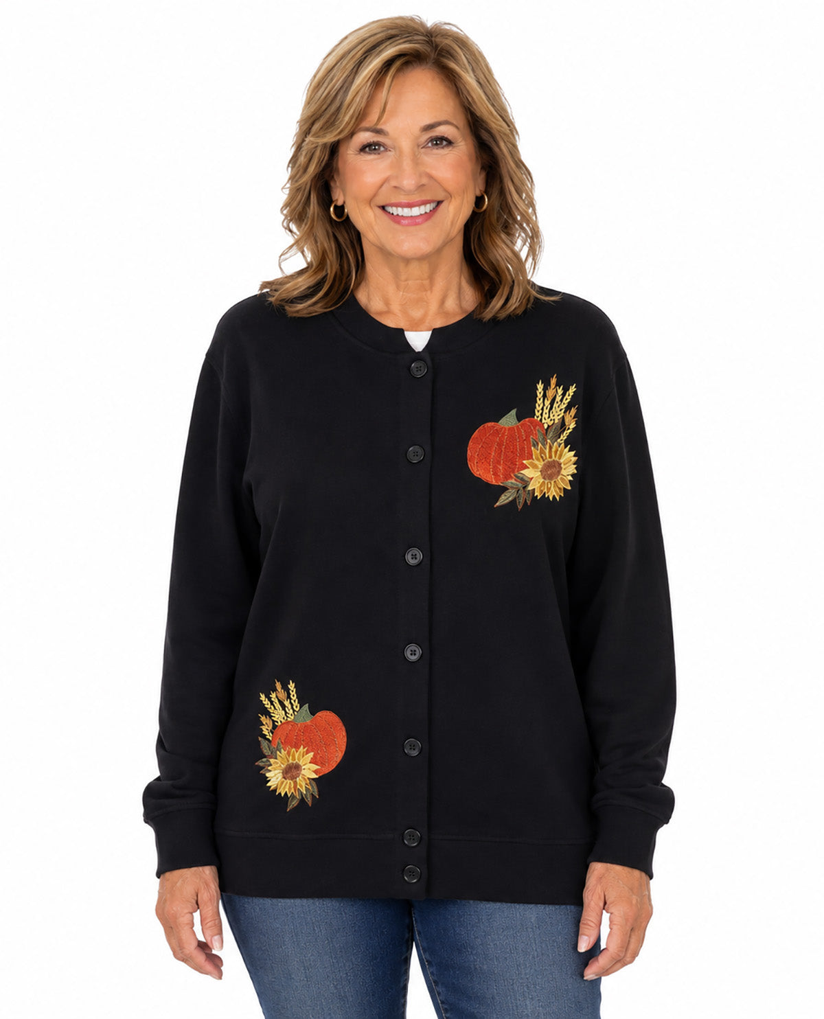 A smiling woman wearing a black button-front cardigan with embroidered autumn motifs, including pumpkins, sunflowers, and wheat stalks, paired with blue jeans against a plain white background.