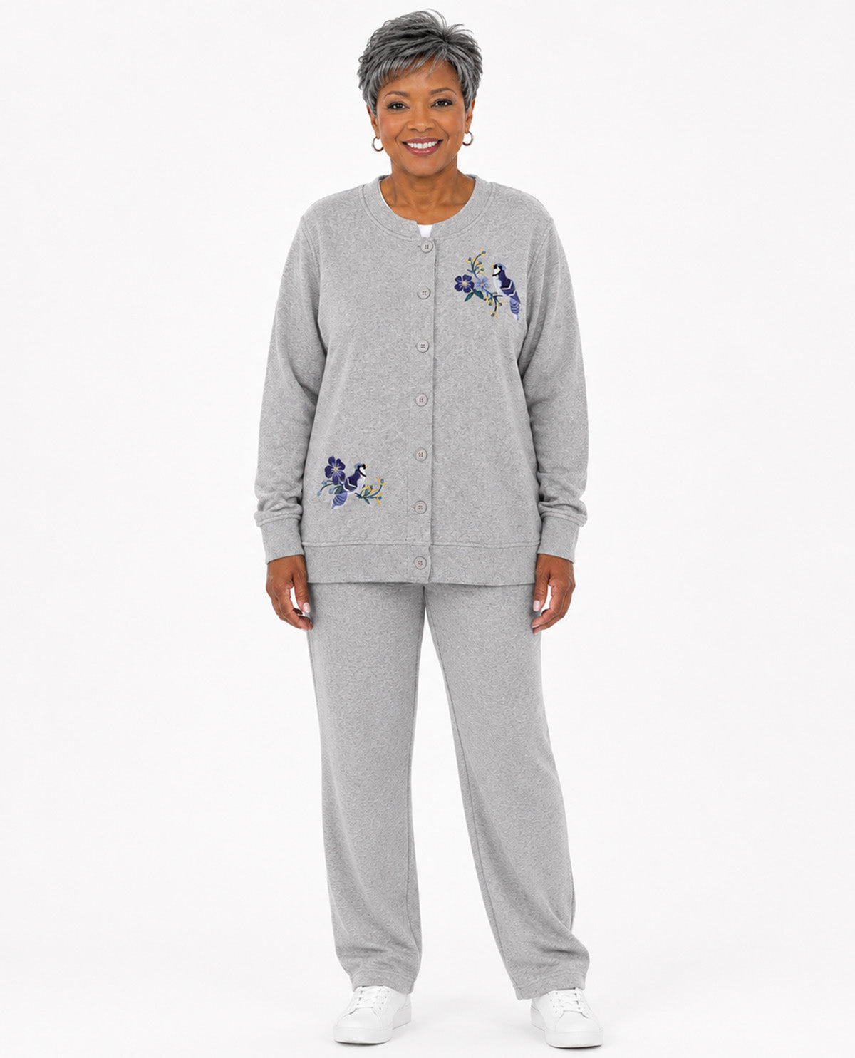 An older woman modeling a light gray heathered two-piece loungewear set, featuring a button-front cardigan with blue and purple floral embroidery on the chest and upper thigh, paired with matching sweatpants, against a plain white studio background.