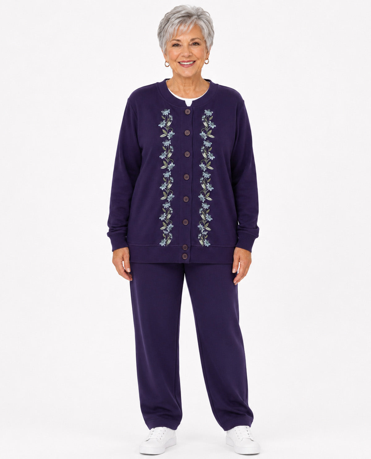 An older woman stands against a light gray studio background wearing a matching two-piece lounge set consisting of a dark purple cardigan and pants with vertical floral embroidery down the front, paired with white sneakers.
