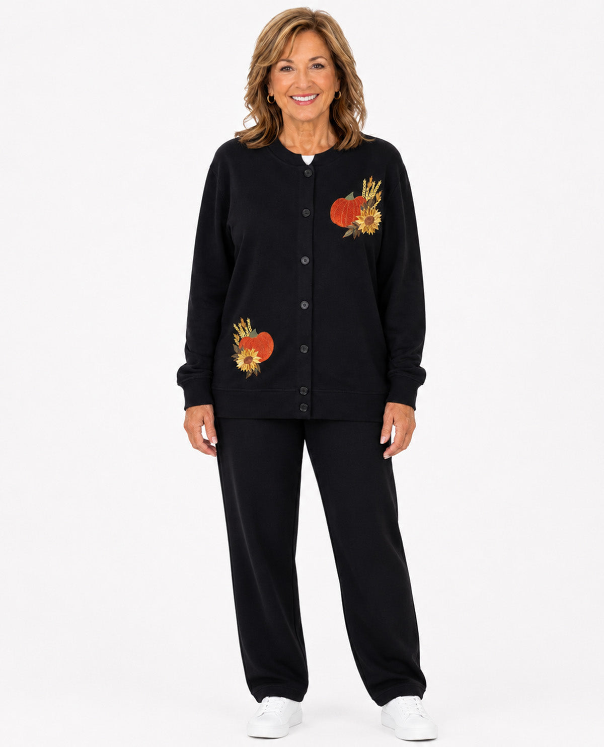 A smiling woman stands against a light gray studio background wearing a black long-sleeve button-front cardigan with embroidered autumn motifs and matching black pants.