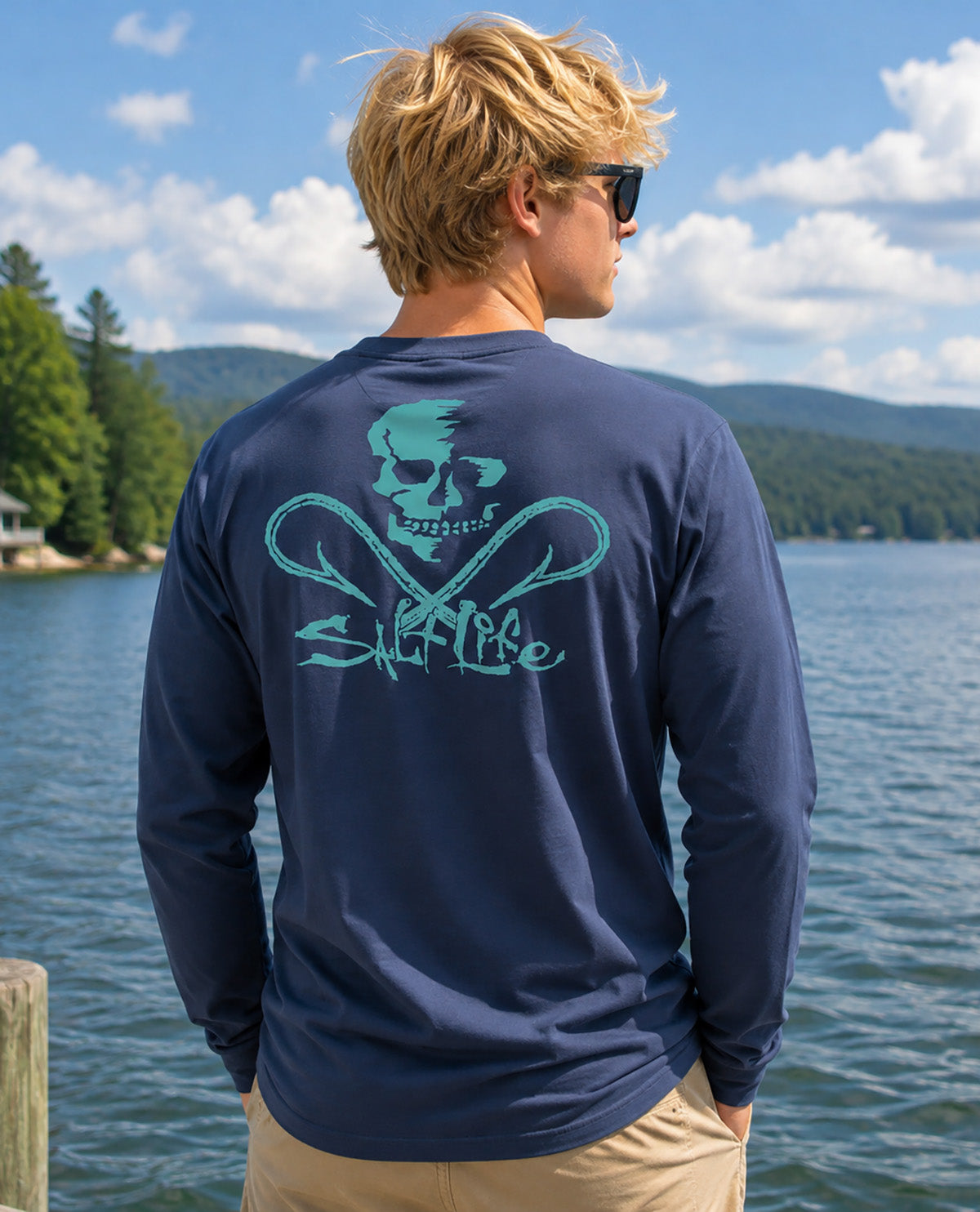 A man standing outdoors by a lake wearing a navy long-sleeve shirt with a large teal skull and crossed-net style graphic on the back, paired with khaki pants and sunglasses.