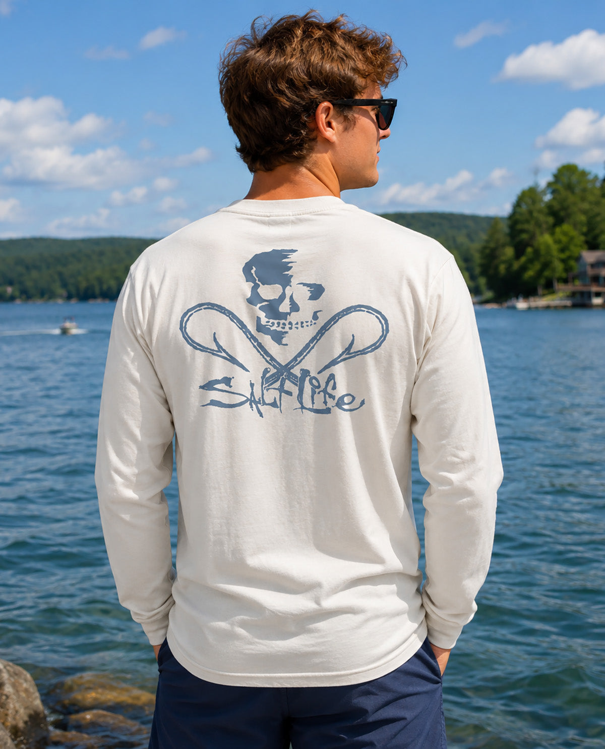 A man wearing a long-sleeve cream t-shirt with a blue skull and crossed anchors graphic on the back, standing by a lake with a scenic background.