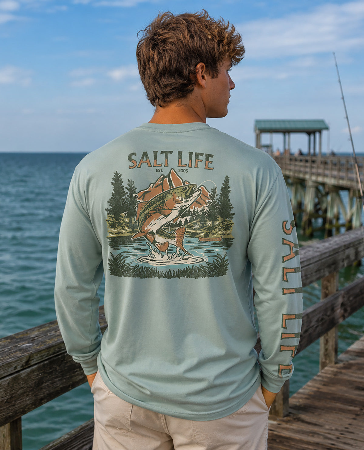 A man viewed from behind wearing a light sage green long-sleeve t-shirt with a large 'Salt Life' fishing graphic on the back and 'SALT LIFE' printed down the sleeve, standing on a wooden pier with the ocean in the background.