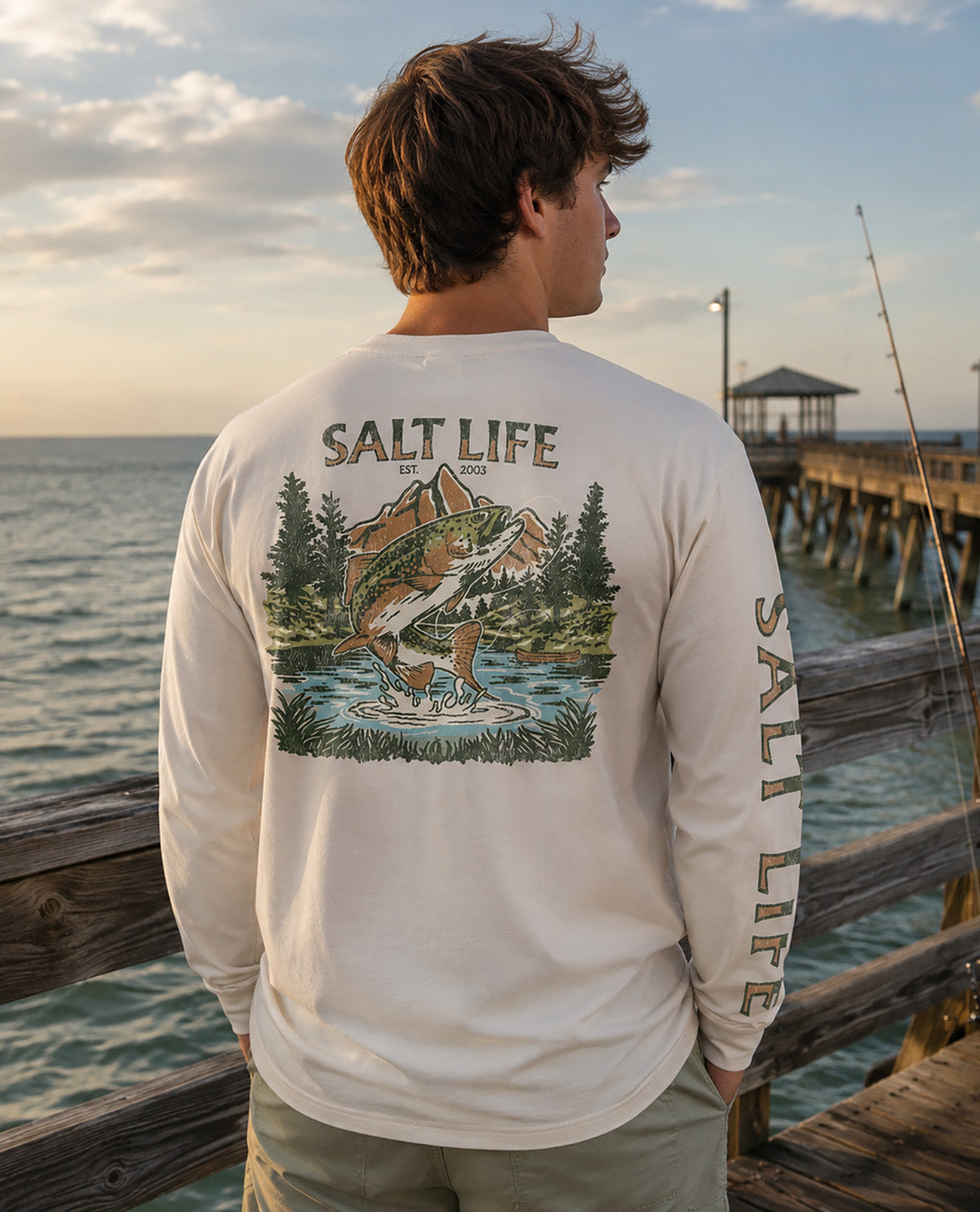 A man stands at a wooden pier overlooking the ocean, wearing a white long-sleeve shirt with a large back graphic of a fish jumping from water, the words 'SALT LIFE' above it, and 'SALT LIFE' printed down one sleeve.