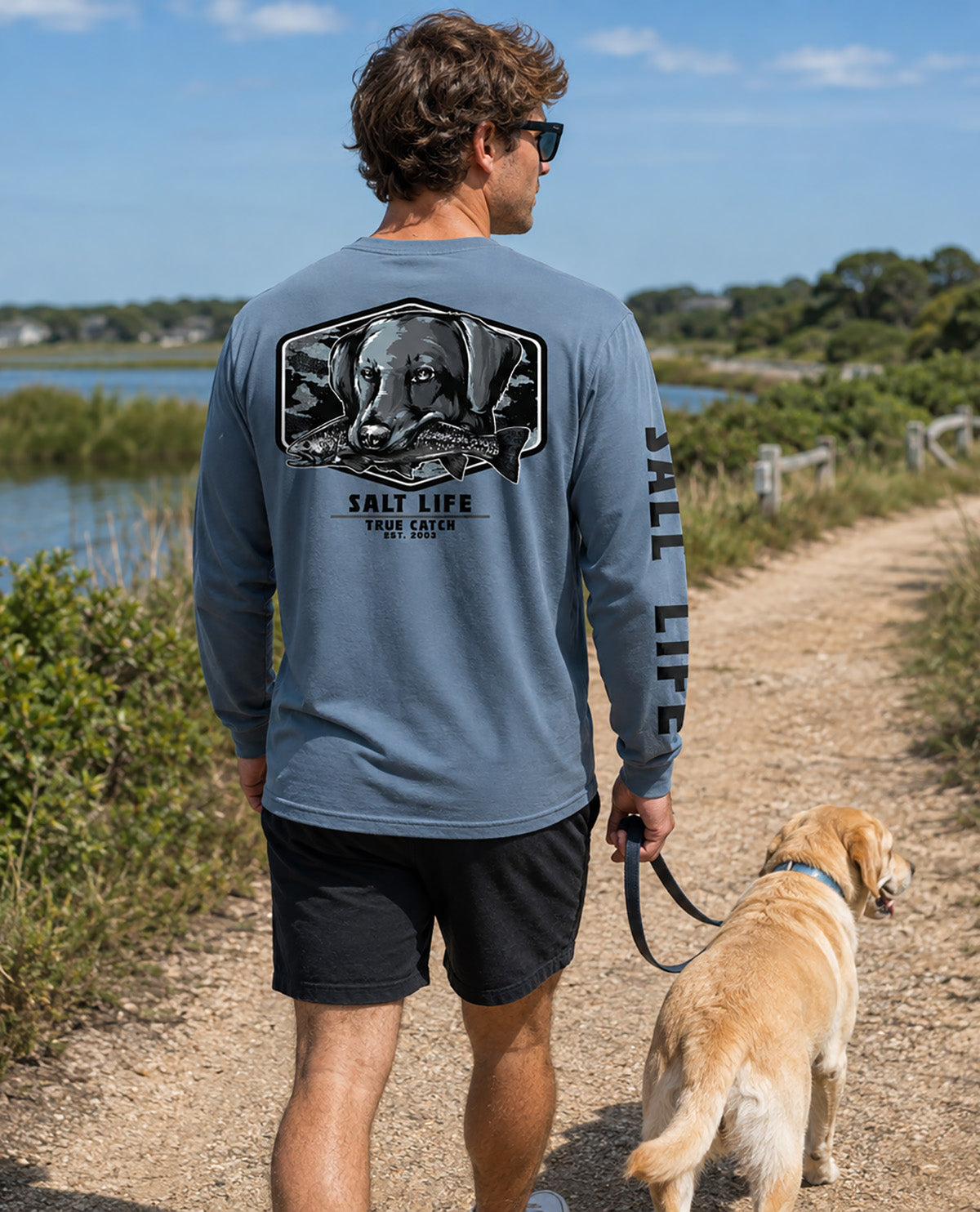 A man wearing a slate blue long-sleeve t-shirt with a large dog graphic on the back and 'SALT LIFE' printed down the sleeve, walking a yellow Labrador retriever on a sandy path outdoors.