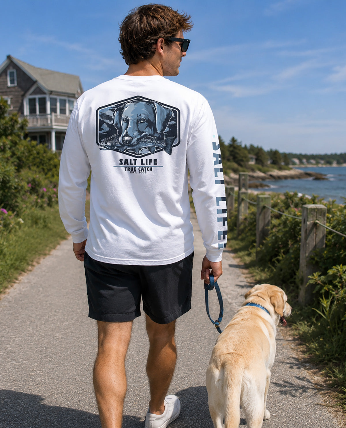 A man wearing a white long-sleeve t-shirt with a large graphic print on the back and 'SALT LIFE' on the sleeve, walking a yellow Labrador retriever on a path outdoors.