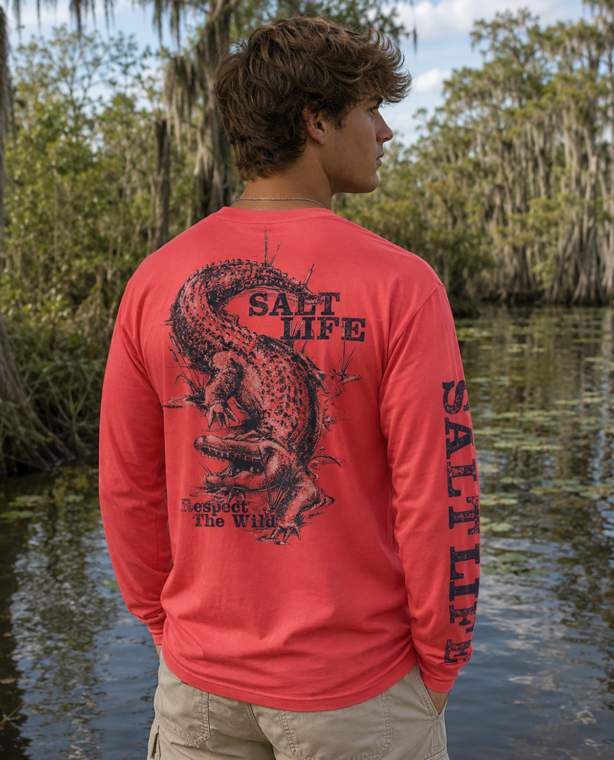 A man standing outdoors by a swampy waterway wearing a coral-red long-sleeve shirt with a large alligator graphic and 'SALT LIFE' text on the back, shown from behind.