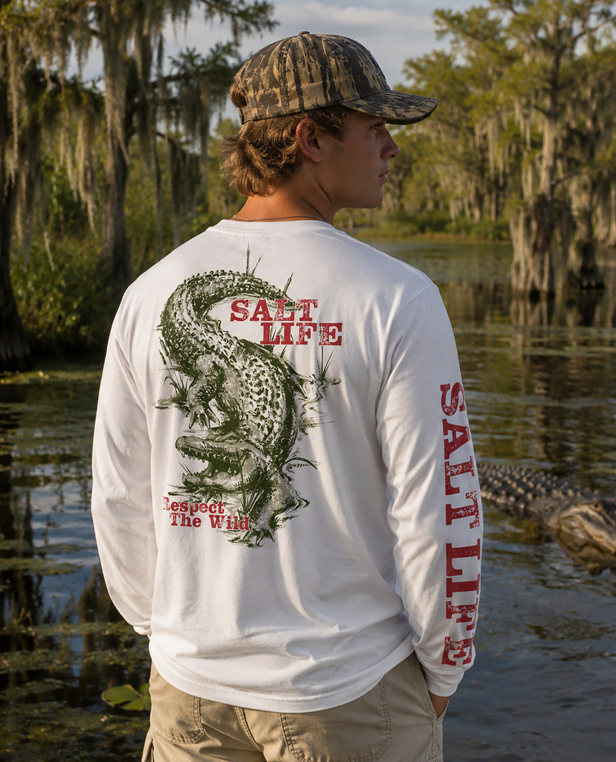A man standing in a swamp wearing a white long-sleeve shirt with a large alligator graphic and red 'SALT LIFE' text on the back and sleeve, paired with a camouflage cap and khaki pants.