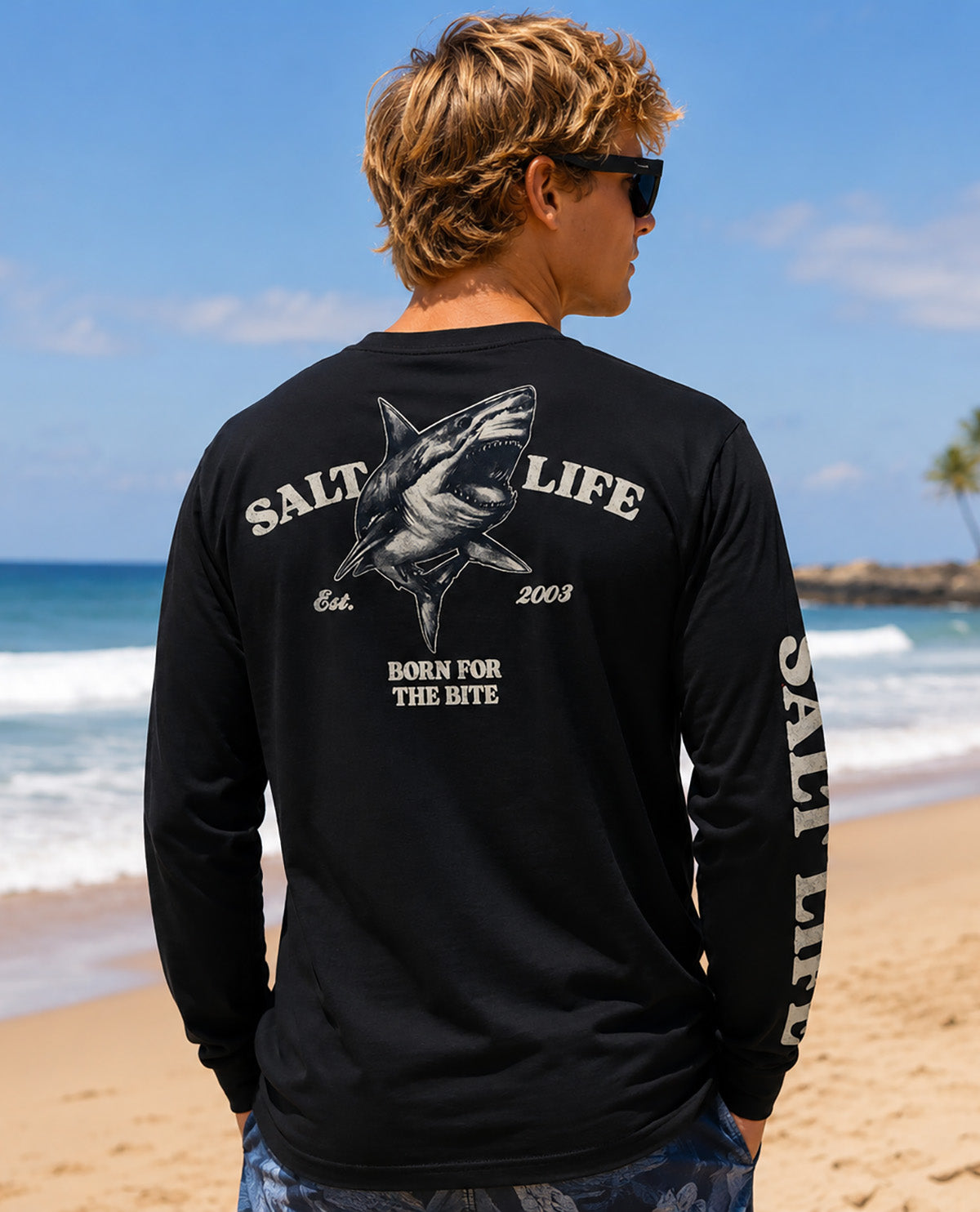 A man standing on a sandy beach facing away from the camera, wearing a black long-sleeve t-shirt with a large shark graphic and 'SALT LIFE' text on the back, and sunglasses.