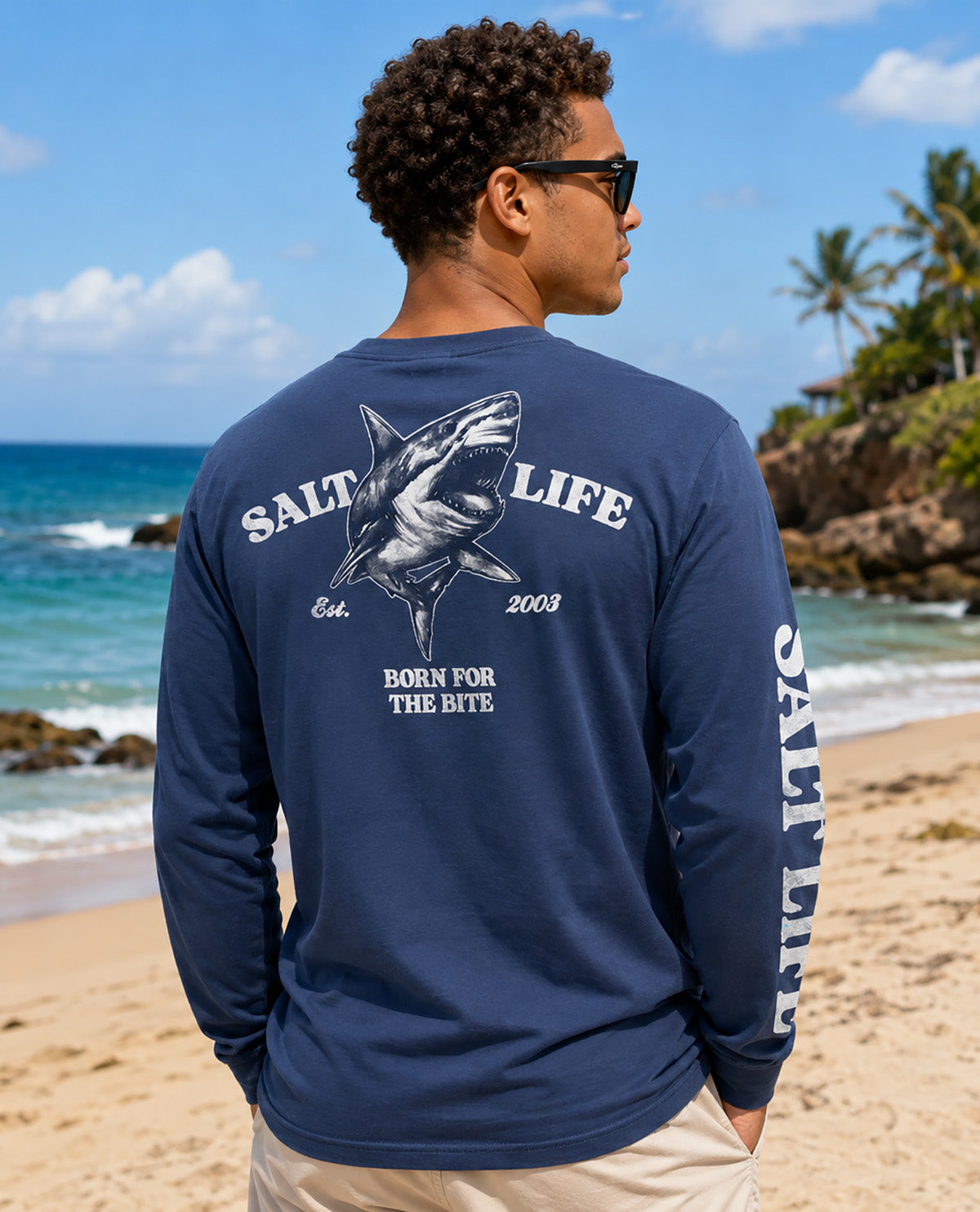 A man wearing a navy blue long-sleeve t-shirt with a large shark graphic and 'SALT LIFE' branding on the back, standing on a sandy beach with the ocean in the background.