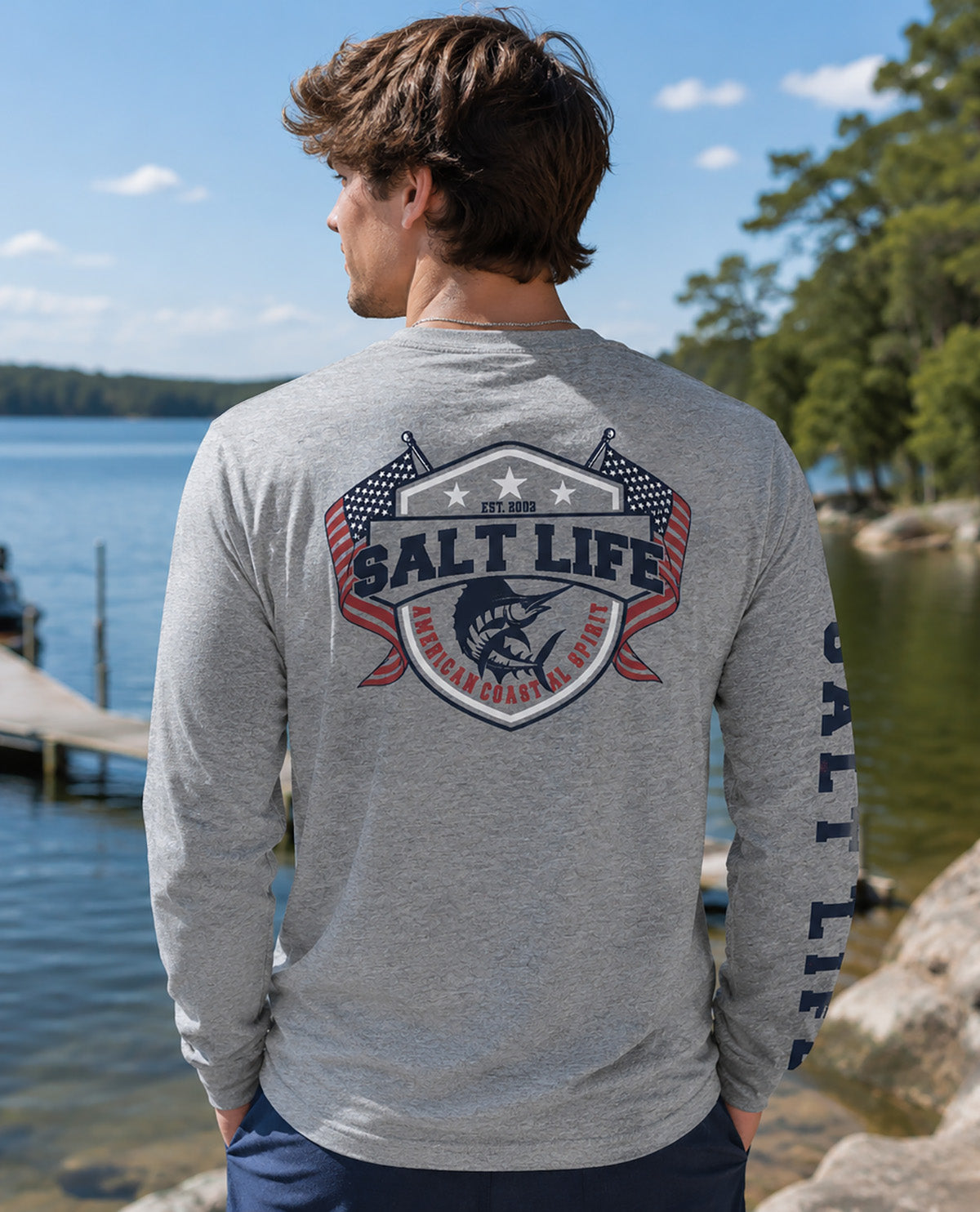A man wearing a heather gray long-sleeve t-shirt with a large 'SALT LIFE' fishing graphic on the back, standing outdoors by a lake with trees in the background.