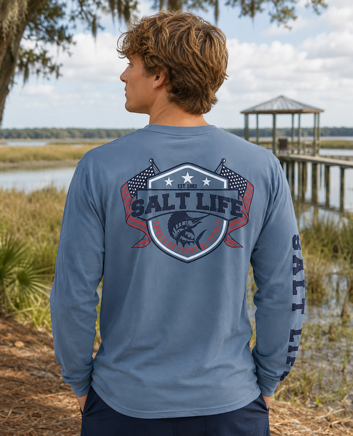 A man wearing a slate blue long-sleeve shirt with a large 'SALT LIFE' fishing graphic on the back, standing outdoors near a lake and wooden dock.
