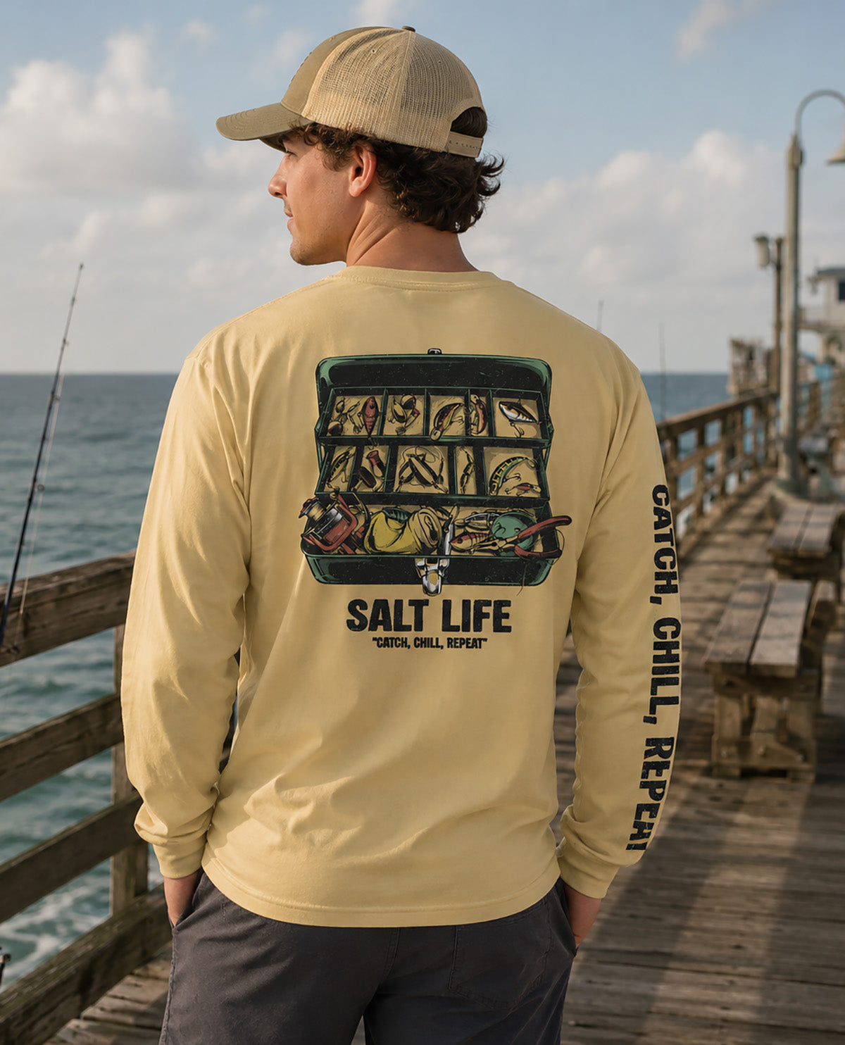 A man wearing a beige baseball cap and a pale yellow long-sleeve graphic shirt stands on a wooden pier with the ocean in the background. The back of the shirt features a large graphic of a fishing tackle box filled with fishing gear and the text 'SALT LIFE' and 'CATCH CHILL, REPEAT.
