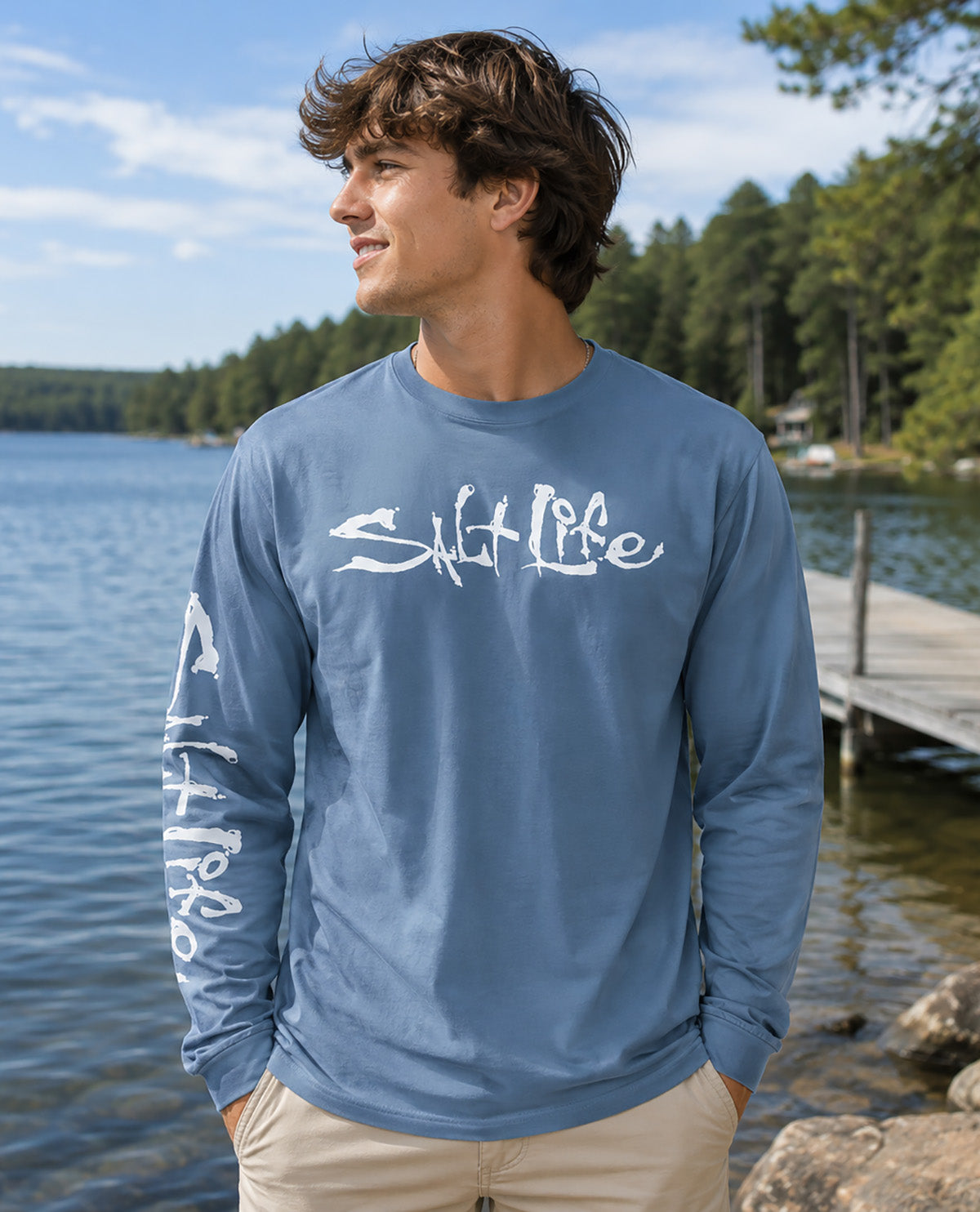 A man wearing a slate blue long-sleeve t-shirt with white 'Salt Life' brand graphics, standing outdoors by a lake with a wooden dock and pine trees in the background.