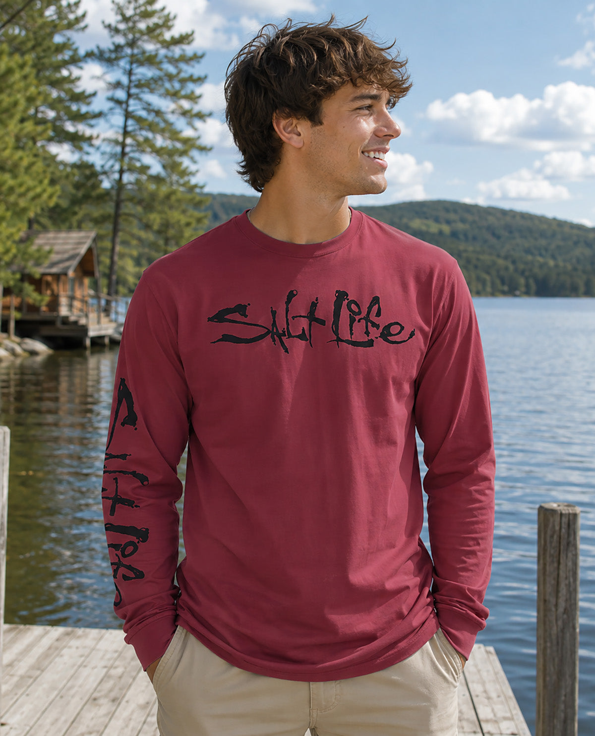 A man wearing a maroon long-sleeve t-shirt with 'Salt Life' printed in black script across the chest and down one sleeve, standing on a wooden dock with a lake, pine trees, and hills in the background.