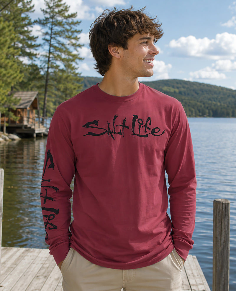 A man wearing a maroon long-sleeve t-shirt with 'Salt Life' printed in black script across the chest and down one sleeve, standing on a wooden dock with a lake, pine trees, and hills in the background.