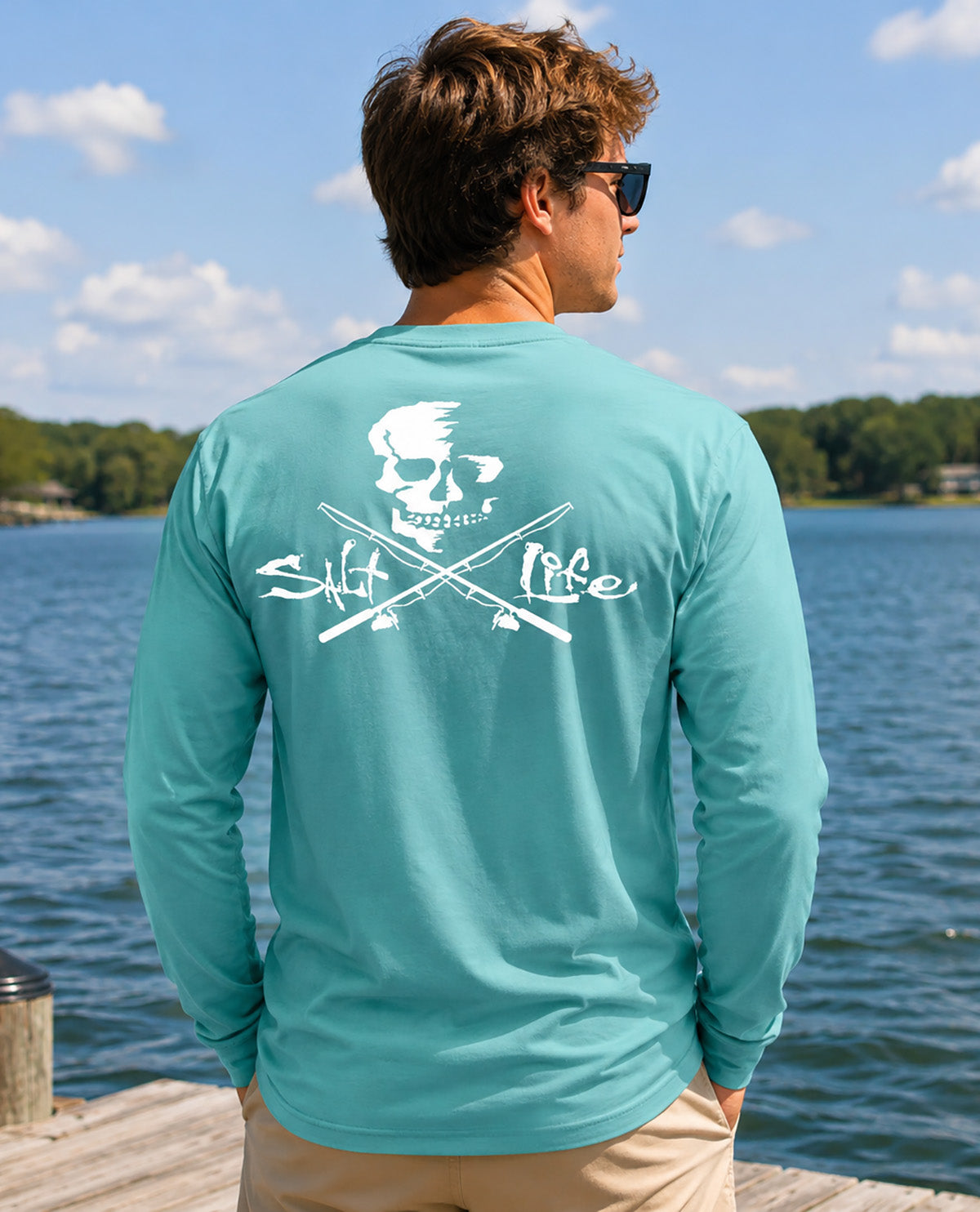 A man viewed from behind wearing a turquoise long-sleeve t-shirt with a white skull and crossed oars graphic on the back, standing on a wooden dock with a lake and trees in the background.