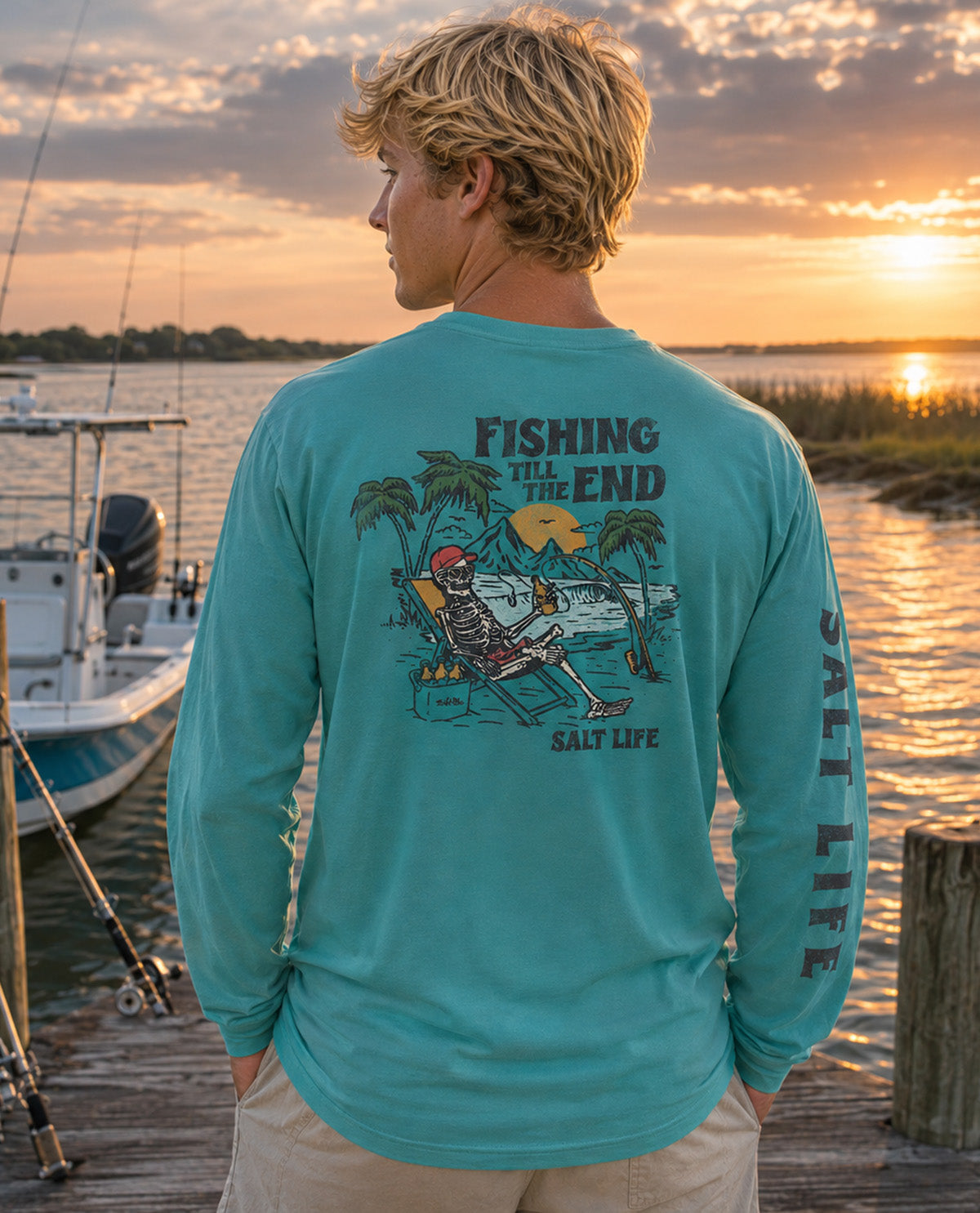 A man stands on a wooden dock at sunset wearing a turquoise long-sleeve graphic shirt. The back of the shirt features a large illustration of a skeleton reclining in a beach chair with palm trees and the text 'FISHING WILL THE END' and 'SALT LIFE'.