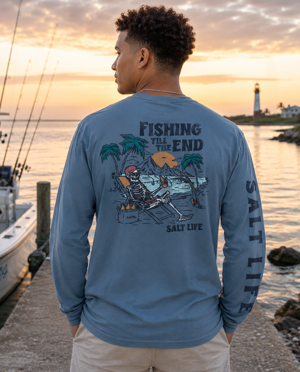 A man stands on a dock at sunset wearing a light blue long-sleeve graphic shirt with a large back print reading 'Fishing Till the End' and 'SALT LIFE,' plus a skeleton lounging by a beach scene. The shirt also has 'SALT LIFE' printed down one sleeve.