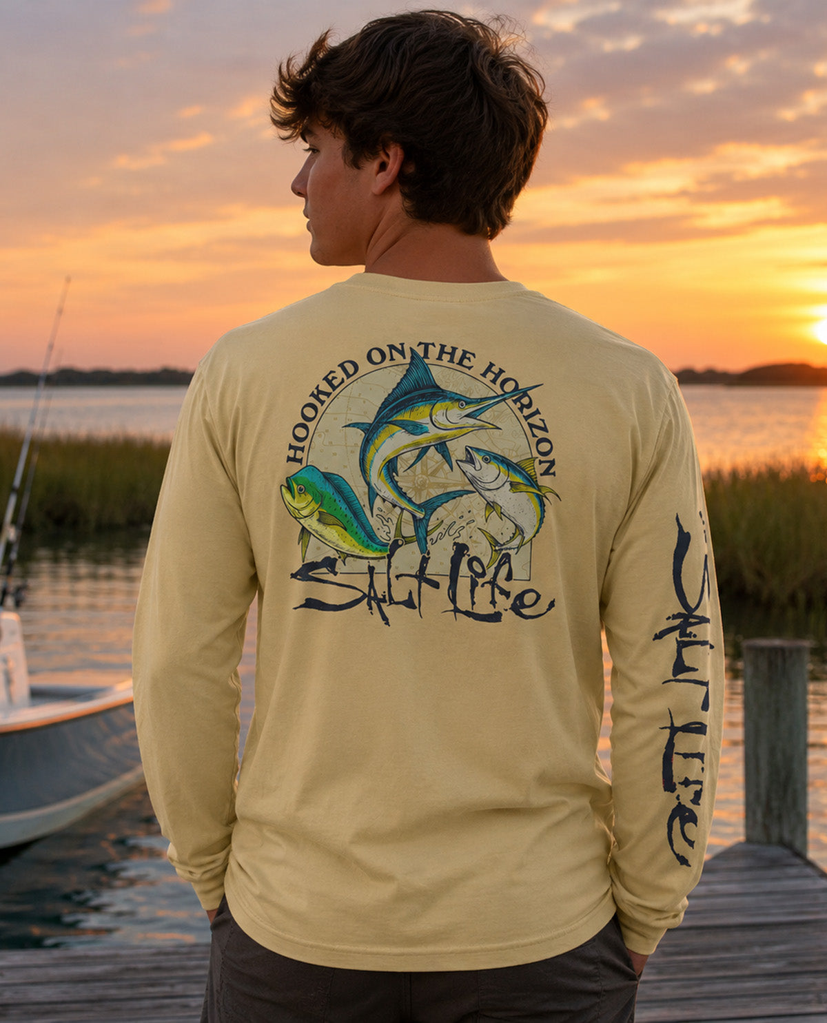 A man stands on a wooden dock at sunset wearing a pale yellow long-sleeve t-shirt with a large back graphic of two fish and the text 'HOOKED ON THE HORIZON Salt Life' on the left sleeve.