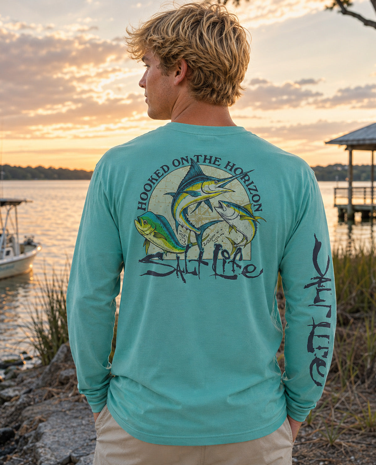 A man stands outdoors by a lake at sunset wearing a turquoise long-sleeve fishing shirt from the back, which features a large graphic of three fish and the text 'HOOKED ON THE HORIZON Salt Life' on the back, with black Japanese characters printed down one sleeve.