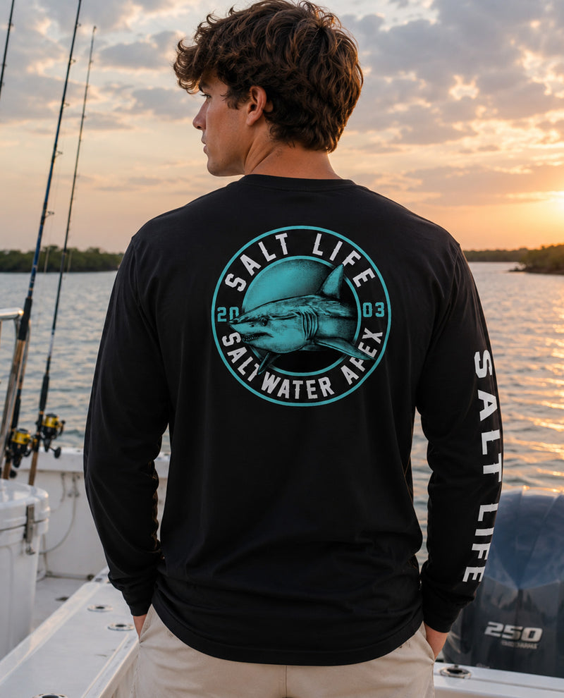 A man on a boat at sunset wearing a black long-sleeve shirt with a large teal and white 'Salt Life' shark graphic on the back and 'Salt Life' printed down one sleeve.