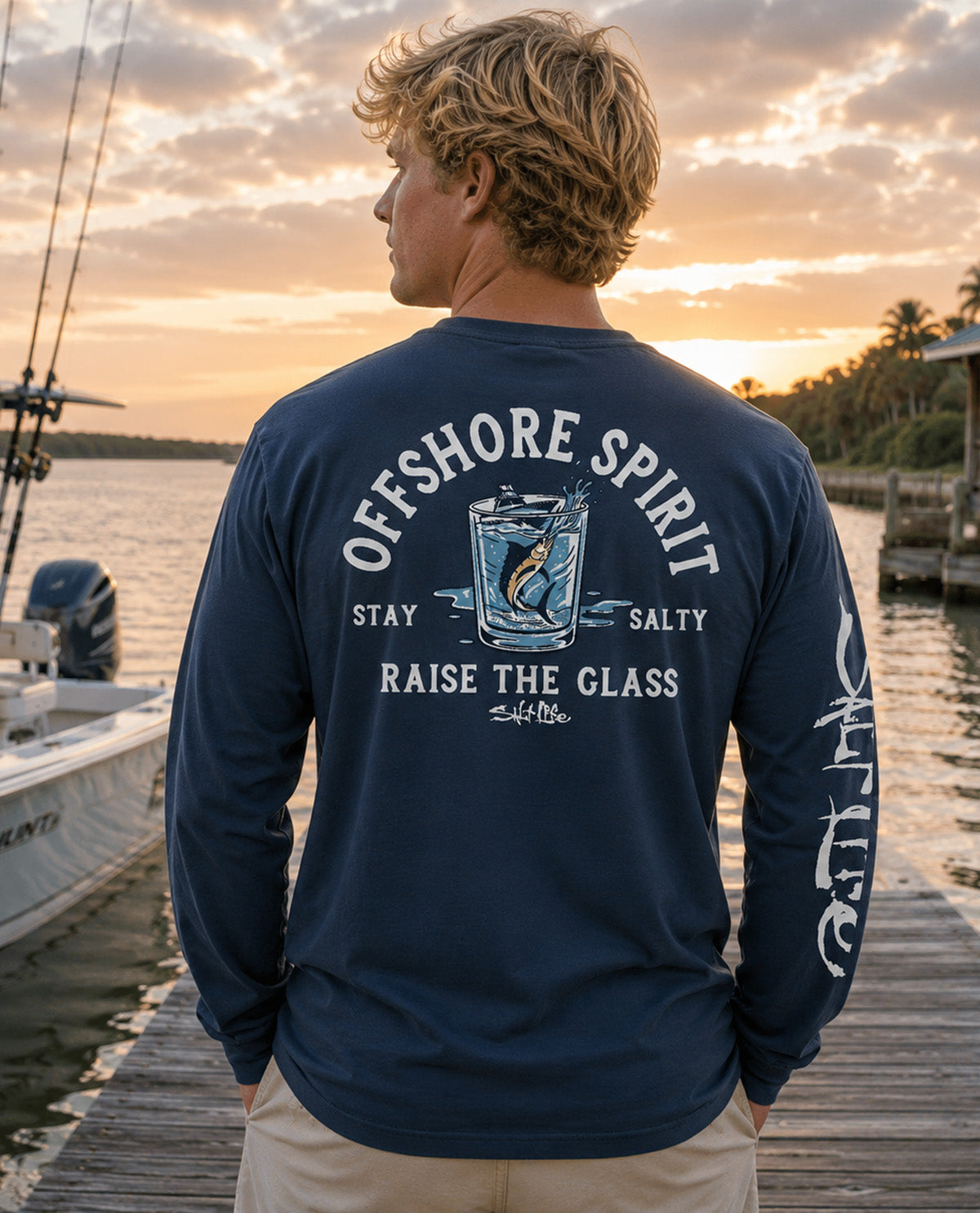 A man standing on a wooden dock at sunset wearing a navy blue long-sleeve shirt with a large back graphic that reads 'OFFSHORE SPIRIT' and 'STAY SALTY RAISE THE GLASS' around a fish-and-glass illustration. The shirt also has white script text down the right sleeve.