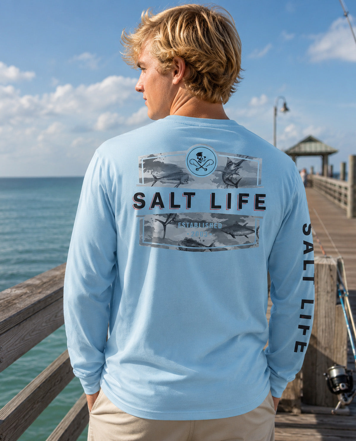 A man wearing a light blue long-sleeve shirt with a large 'SALT LIFE' fish graphic on the back, standing on a wooden pier with the ocean and sky in the background.