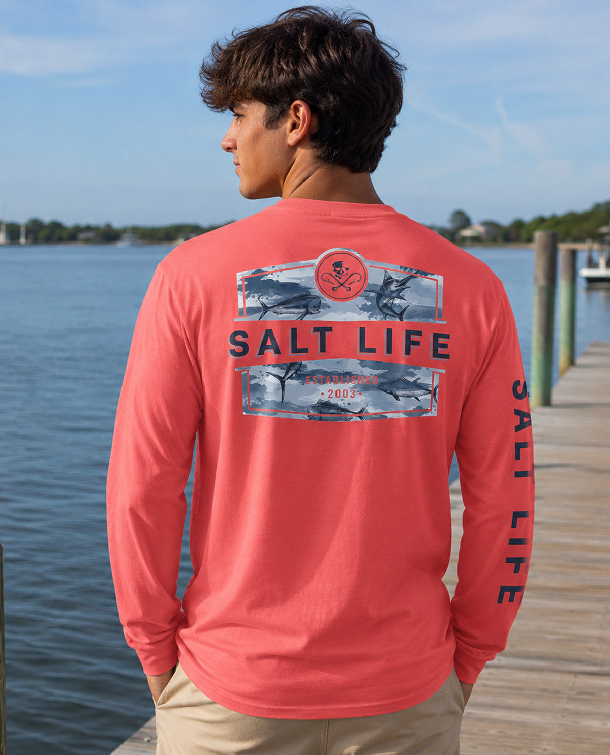 A man wearing a coral pink long-sleeve t-shirt with a 'SALT LIFE' graphic on the back, standing on a wooden dock with water and a distant shoreline in the background.