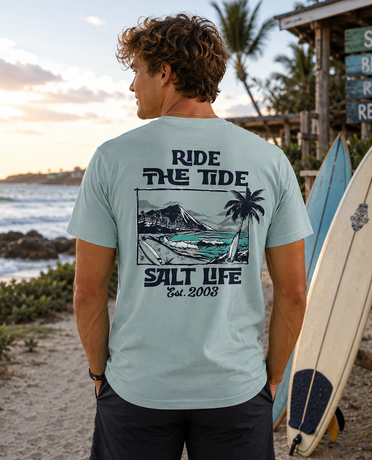 Male model wearing a lt blue salt life tee with saying "RIDE THE TIDE" on back of shirt with oceanfront background