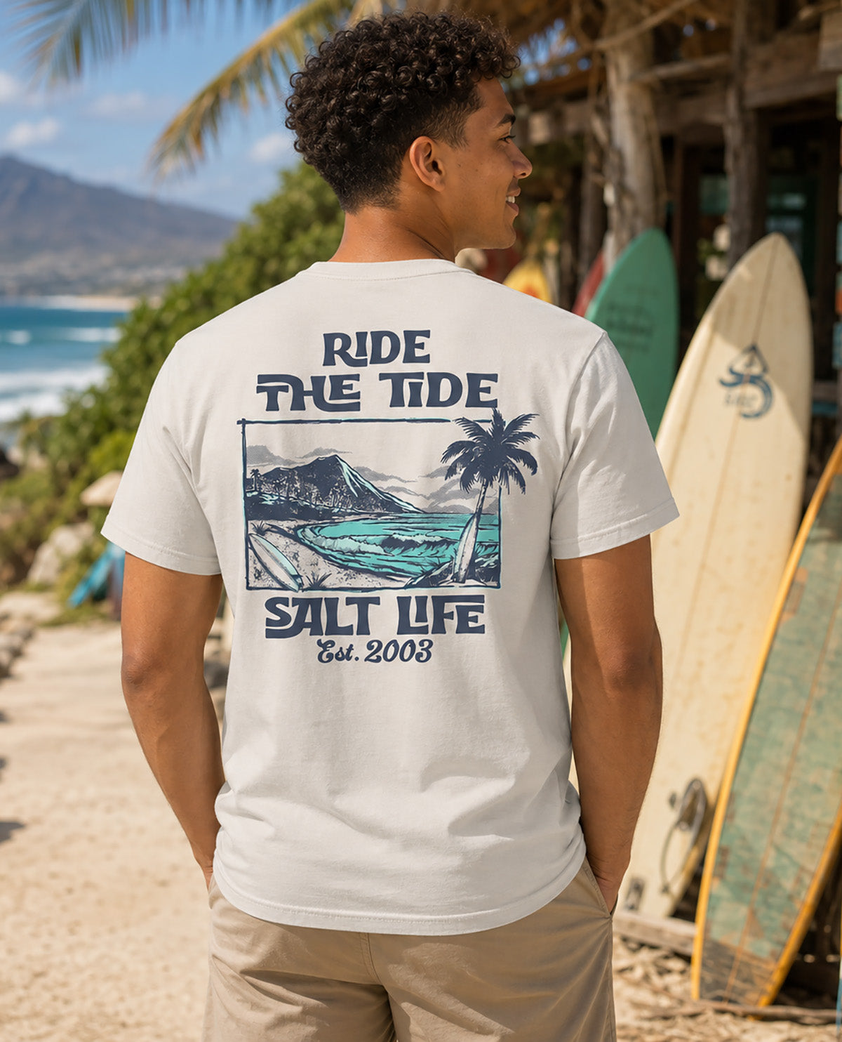 A man wearing a white t-shirt with a blue graphic print on the back, standing on a beach with surfboards and a building in the background. The text on the shirt reads 'RIDE THE TIDE', 'SALT LIFE', and 'Est.