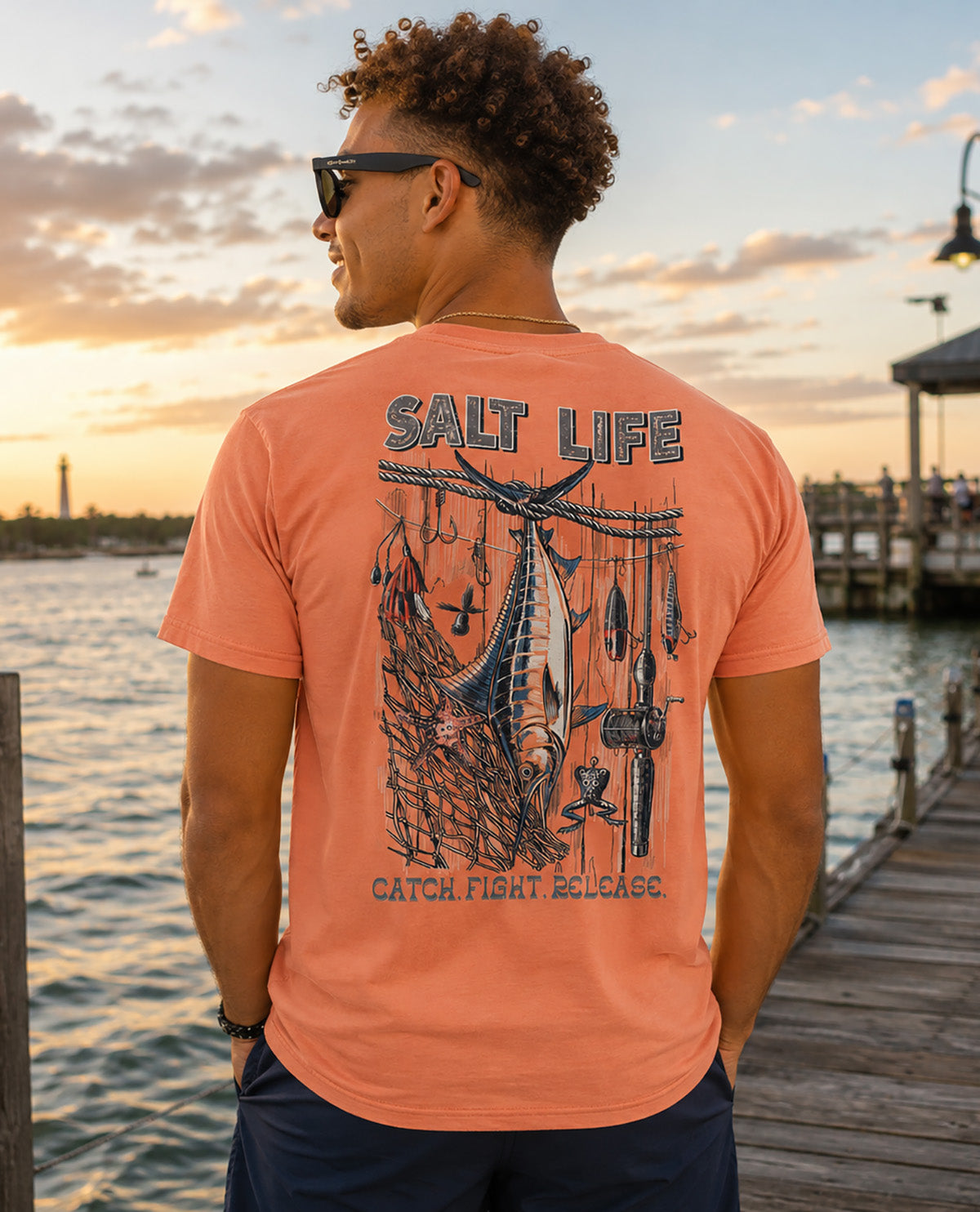 A man wearing a coral orange t-shirt with a large navy blue 'SALT LIFE' fishing graphic on the back, standing on a wooden dock at sunset.