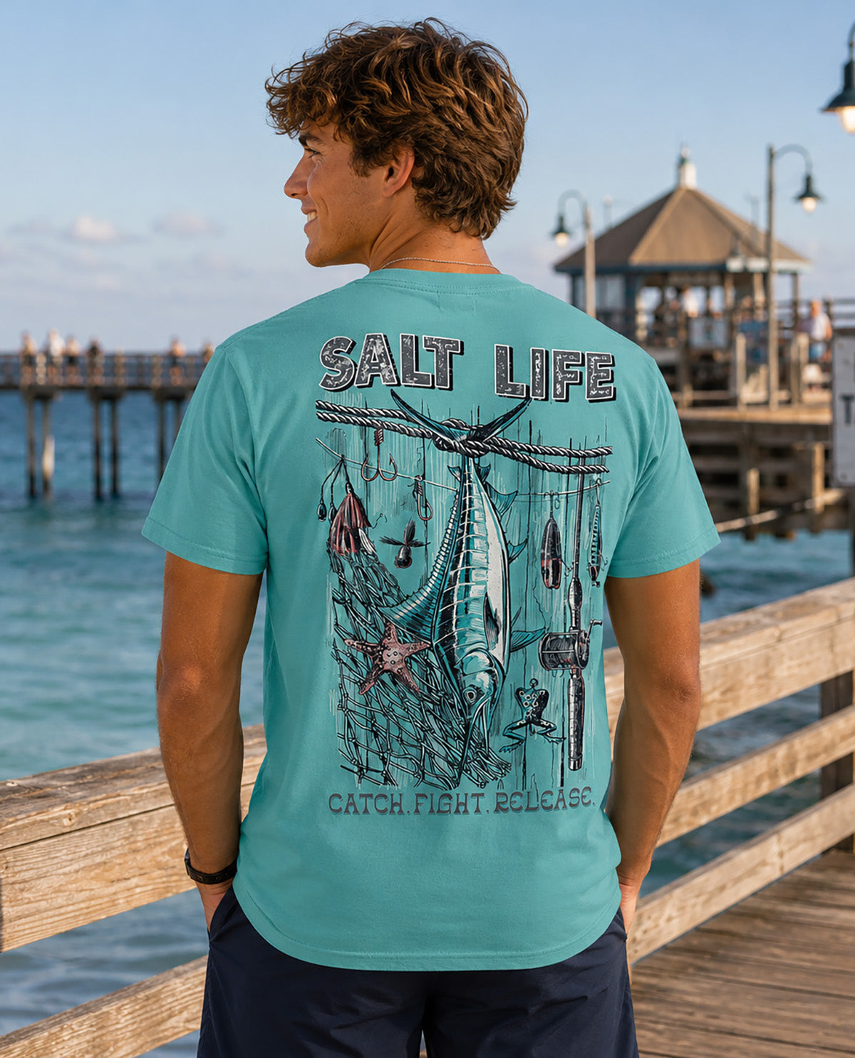 A man standing on a wooden pier at a seaside location, wearing a turquoise t-shirt with a large back graphic that reads 'SALT LIFE' and 'CATCH, FIGHT, RELEASE'.