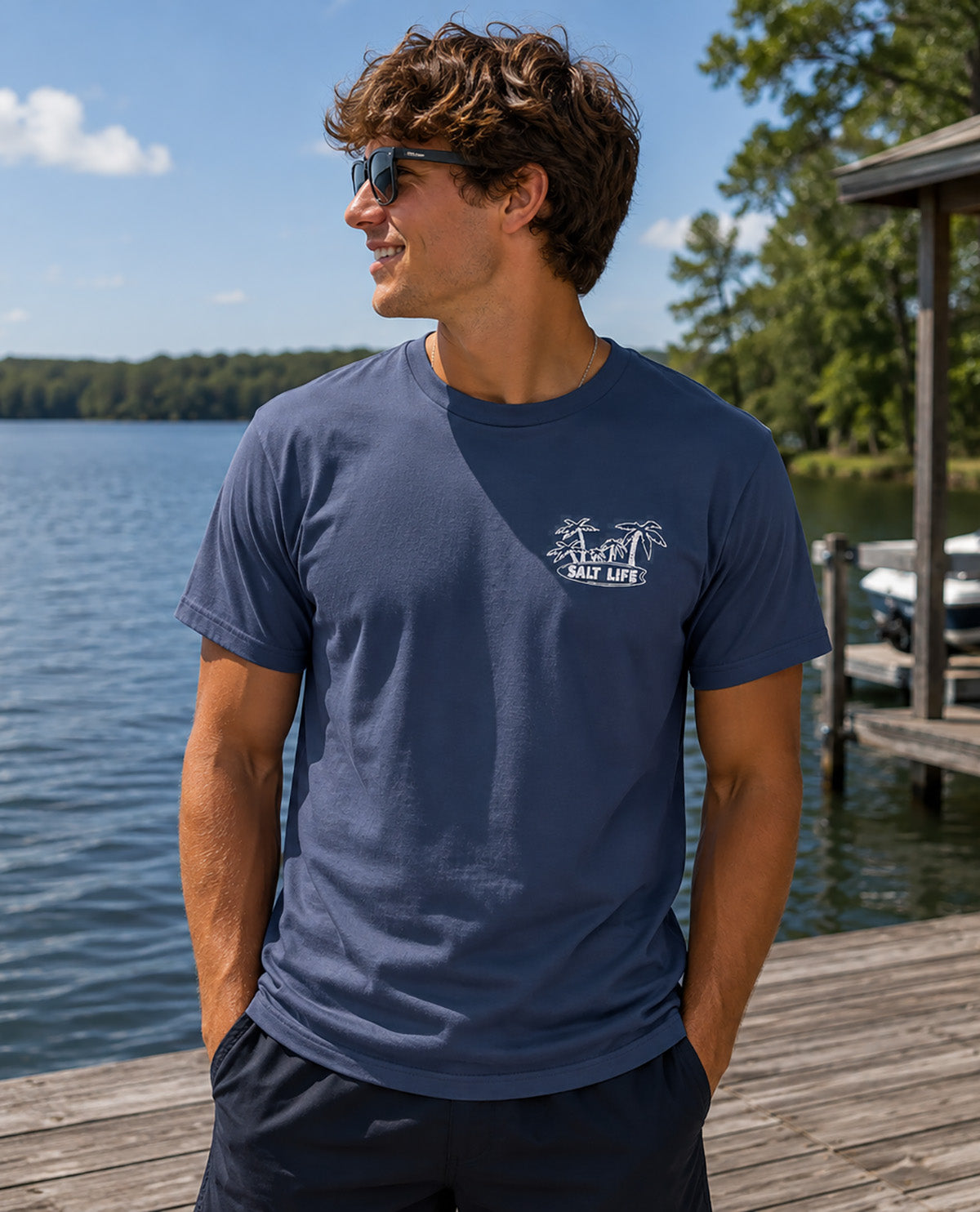 A man wearing a navy blue crewneck t-shirt with a white 'Salt Life' palm tree logo on the left chest, standing on a wooden dock next to a lake.