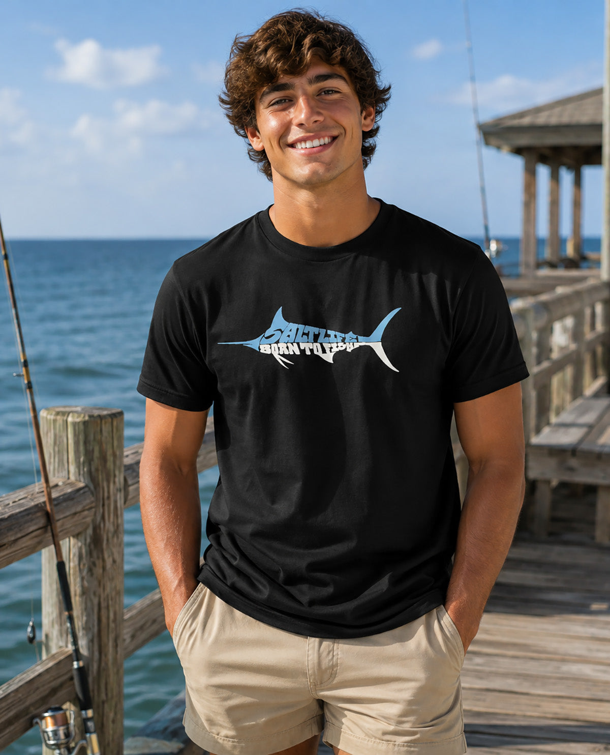 A smiling man wearing a black t-shirt with a blue and white marlin graphic and the text 'BORN TO FISH' stands on a wooden dock with the ocean in the background.