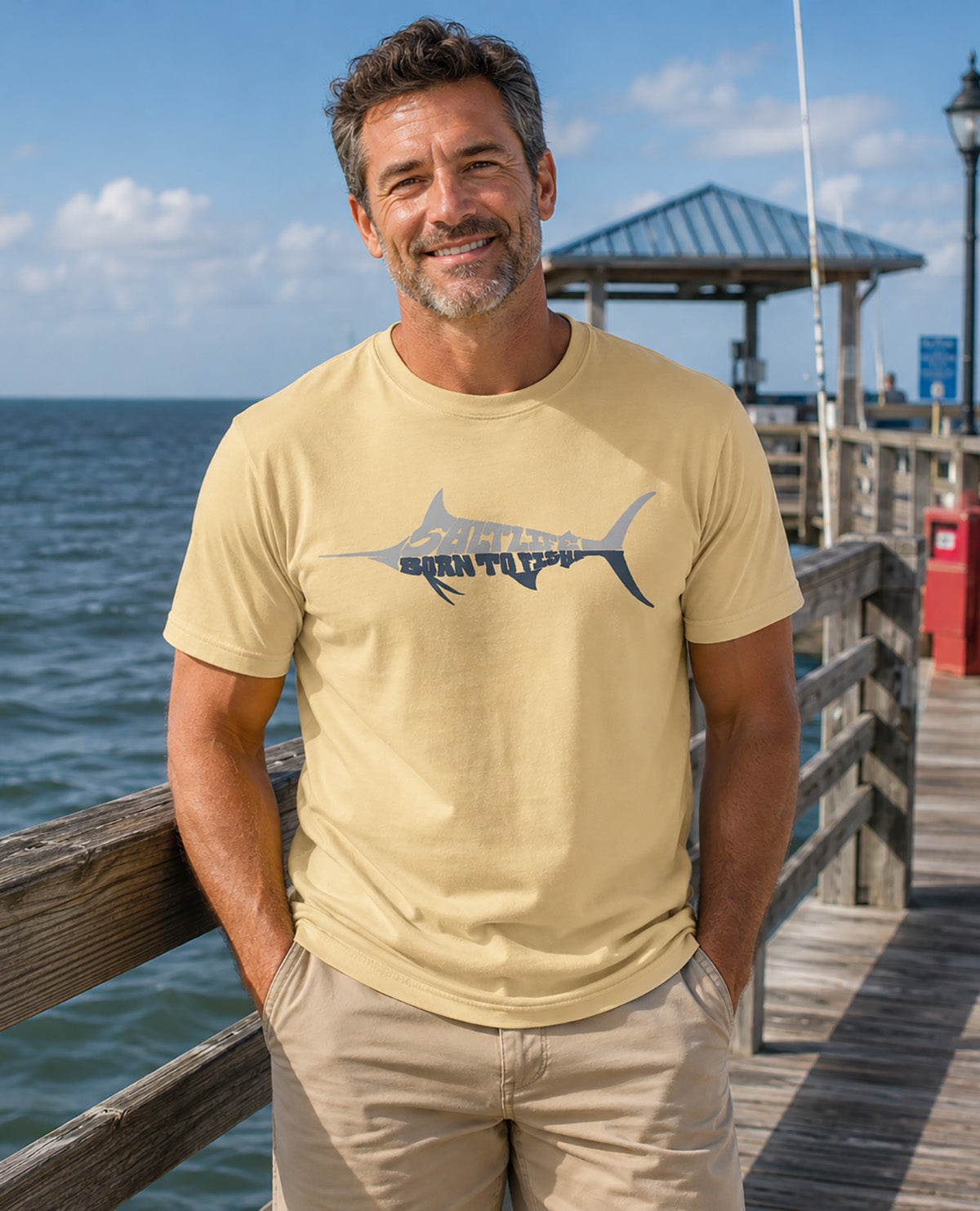 A smiling man wearing a light beige t-shirt with a dark blue marlin graphic that reads 'BORN TO FISH' stands on a wooden pier with the ocean and a pier structure in the background.
