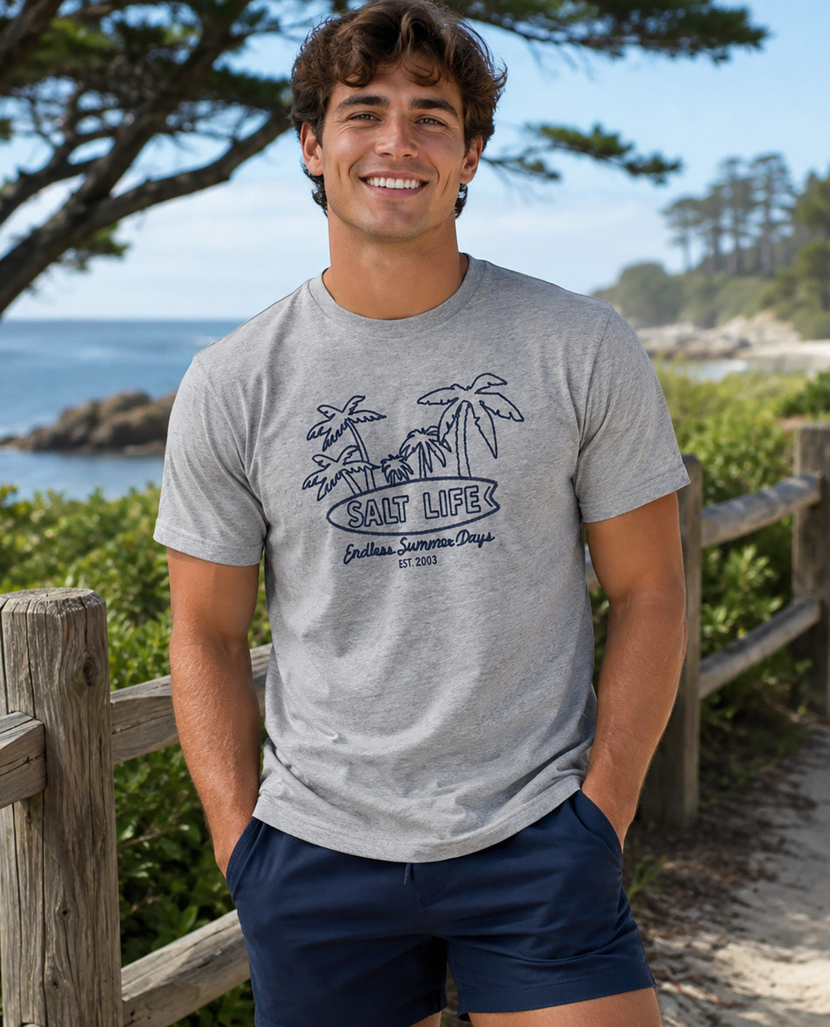 A smiling man wearing a heather gray graphic t-shirt and navy blue shorts, standing outdoors on a beachside path with a wooden fence, greenery, and the ocean in the background.