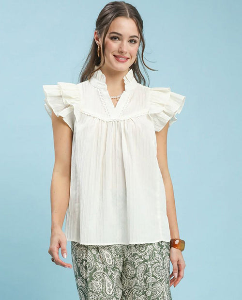 A woman modeling a white ruffled blouse with a pleated bodice and patterned pants against a light blue studio background.