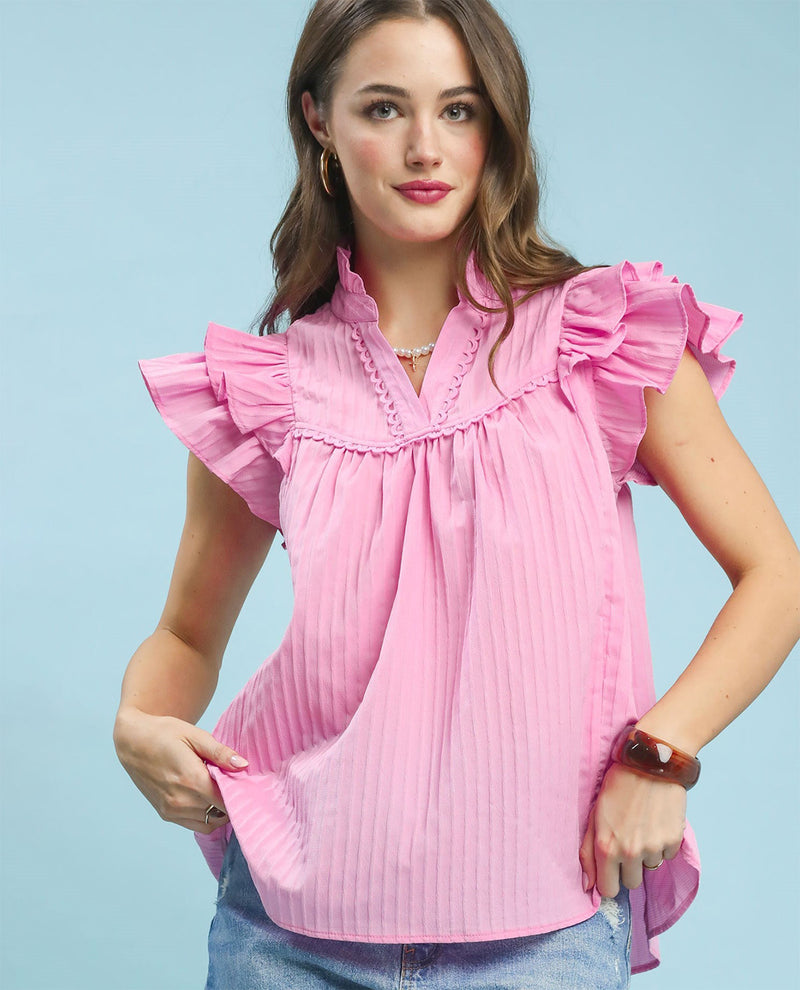 A woman wearing a light pink pleated blouse with ruffled cap sleeves and a ruffled collar, paired with blue jeans, against a solid light blue background.