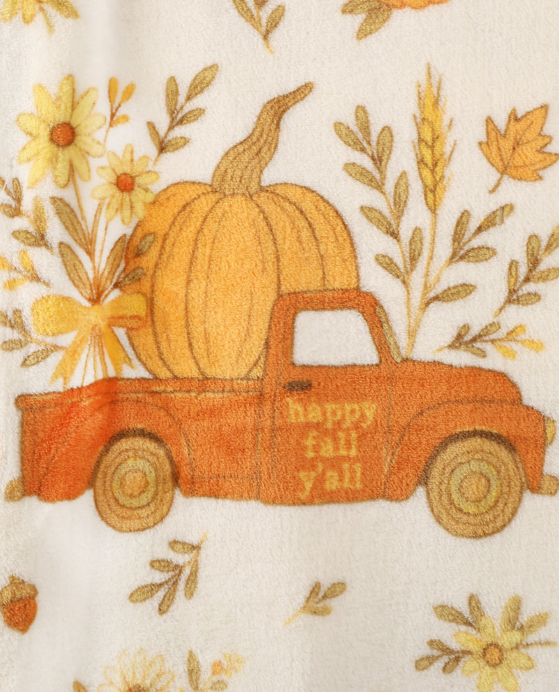 A close-up of a fabric featuring an autumn-themed print with a vintage orange pickup truck carrying a large yellow-orange pumpkin, surrounded by yellow flowers, wheat stalks, leaves, and acorns on an off-white background. The side of the truck reads 'happy fall y'all'.