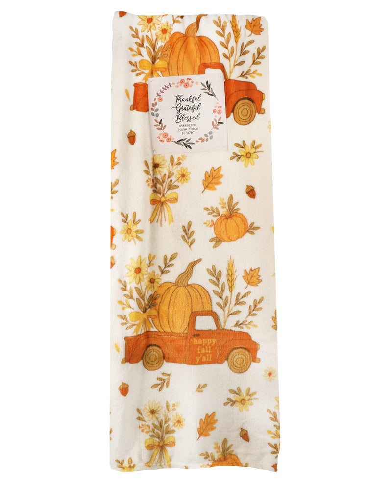 A vertical fabric item, likely a tea towel or kitchen towel, featuring a fall harvest print with orange pickup trucks carrying pumpkins, yellow flowers, and autumn leaves on an off-white background. A white paper label with 'Thankful Grateful Supply' is attached near the top.